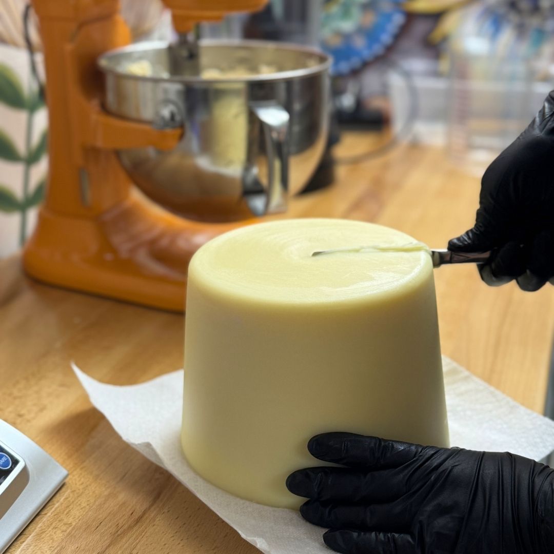 Person wearing black gloves cutting a brick of grass fed tallow on a wooden table with a stand mixer in the background.